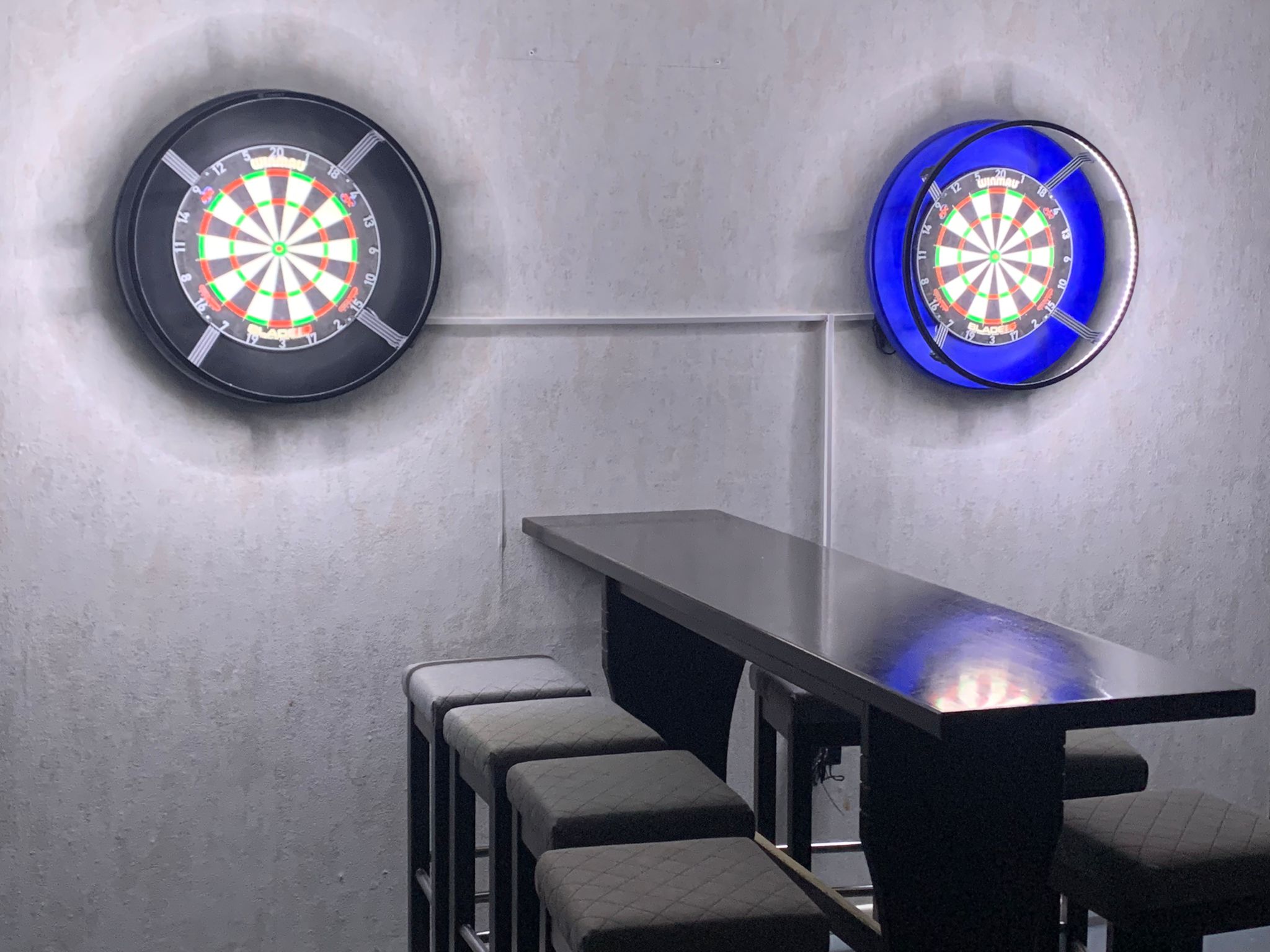 eine blaue und eine schwarze Dartscheibe hängen an der Wand. Davor steht ein langer Tisch mit Hochstühlen.